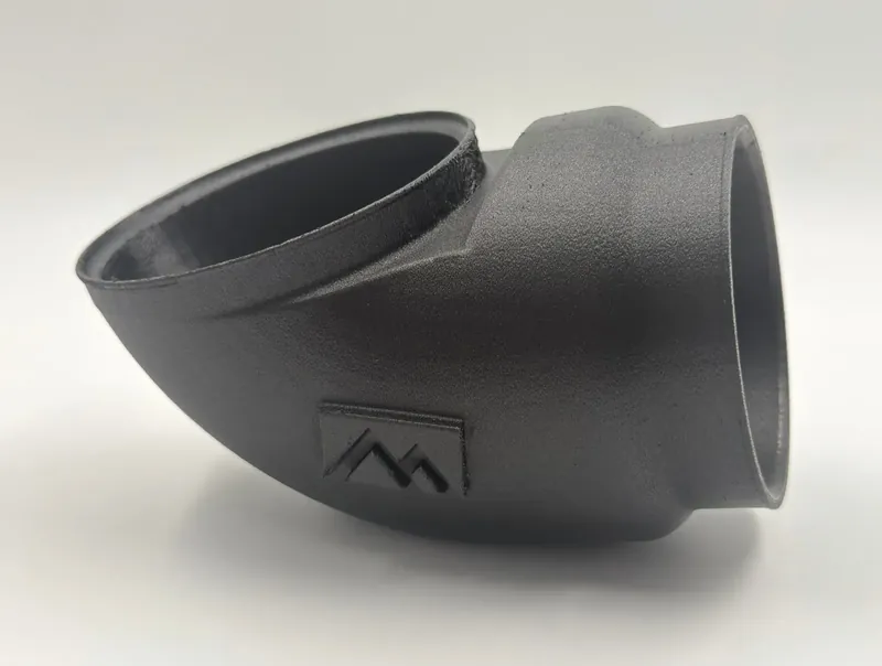 Black air intake elbow 3D printed in carbon-filled PPA: a curved tube with a round opening at each end, a smooth satin surface, and a small raised mountain logo on its side.