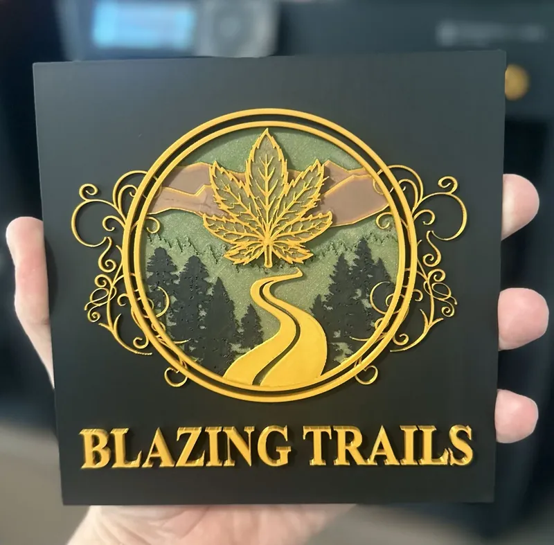 Square black 3D printed plaque held in a hand, with a raised gold circular emblem: a cannabis leaf over a winding road through pine trees and mountains in green and tan, gold scrollwork on both sides, and raised gold lettering reading 'Blazing Trails' below.