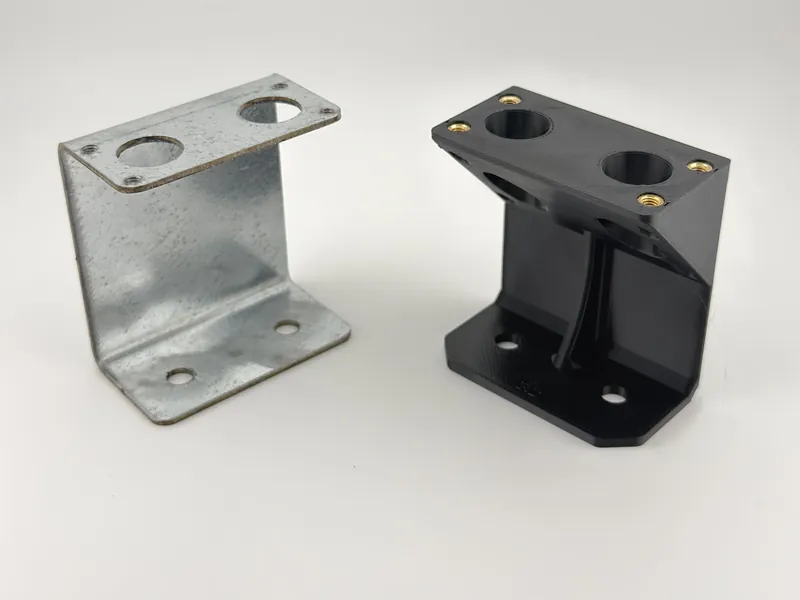 A galvanized steel bracket next to its redesigned black 3D printed alternative, each with two large holes in the top flange and mounting holes in the base.