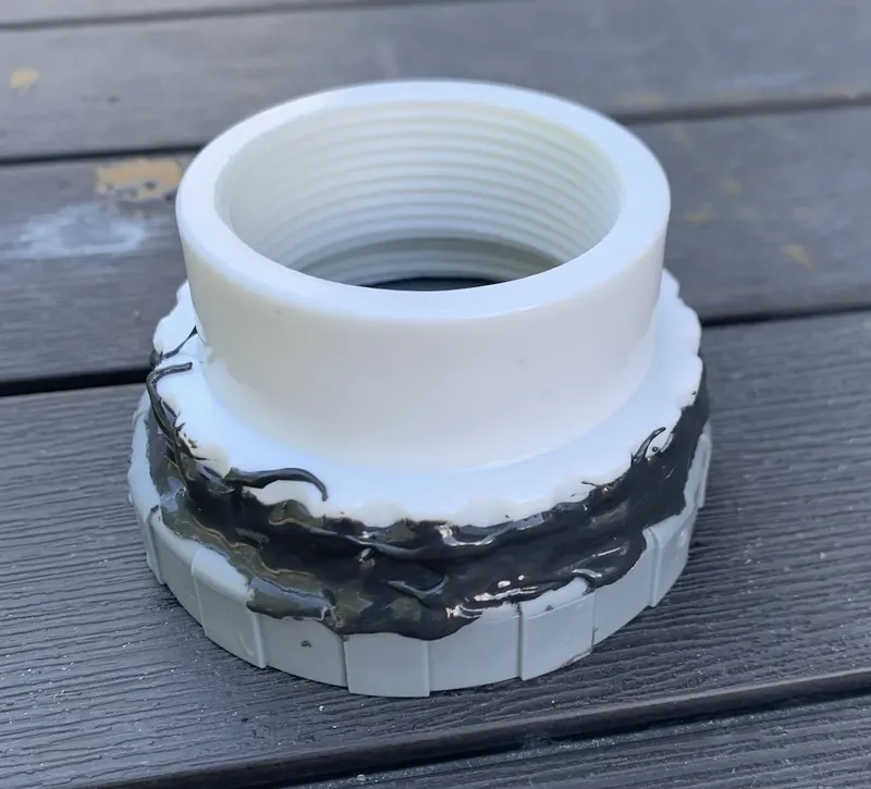 The original fitting, glued together: a white neck threaded on the inside, set into a gray ribbed ring, with black sealant smeared over the joint, on a wooden deck.