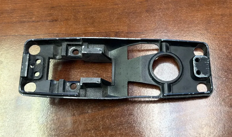 The original cast gate latch part we scanned, seen from its hollow underside: black paint chipped to bare metal along the rim, a large round bore in a raised ring, a rectangular opening between two screw bosses, and four corner holes, on a wood table.