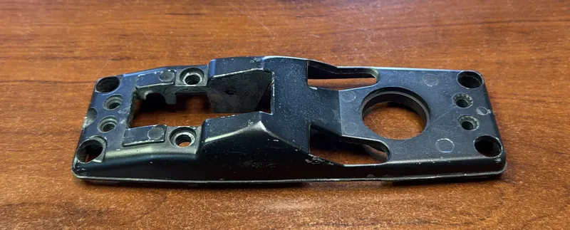 The original cast gate latch part we scanned, seen from its top face: black paint worn to bare metal at the edges, a large round bore, a raised angled section beside a rectangular opening, and four corner holes, on a wood table.