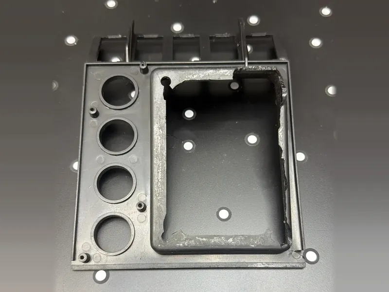 The original panel we scanned, seen from its back face: four round holes with raised rims, small screw bosses, a rectangular frame around the large opening with one broken corner, and five box-like housings along the top edge, on a black table with white dots.