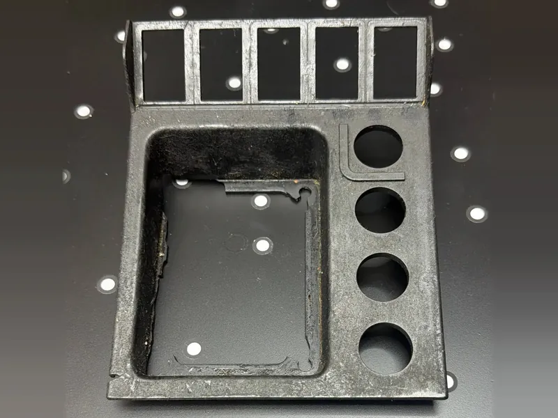 The original panel we scanned, seen from its front face: a row of five rectangular openings along the top edge, a large rounded pocket, and four round holes down the right side, with worn, scuffed surfaces, on a black table with white dots.