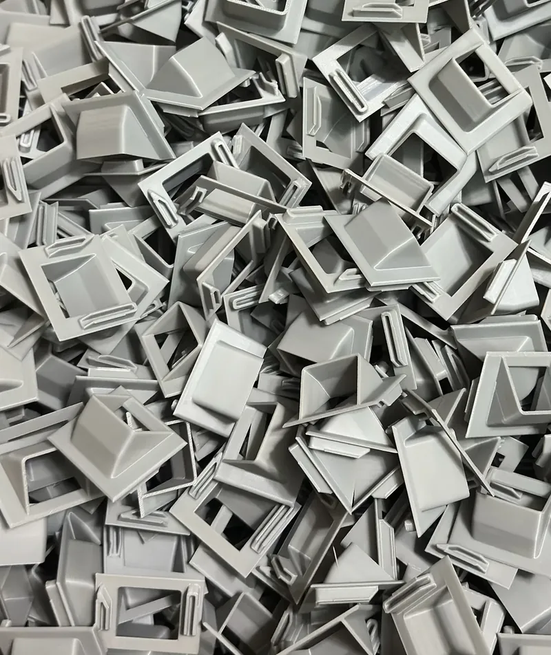 A heap of gray 3D printed clip parts, each a square frame with a raised wedge and small tabs.