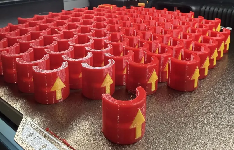 A printer plate covered in rows of red 3D printed clips, each with a raised yellow arrow, with one finished clip standing in front.
