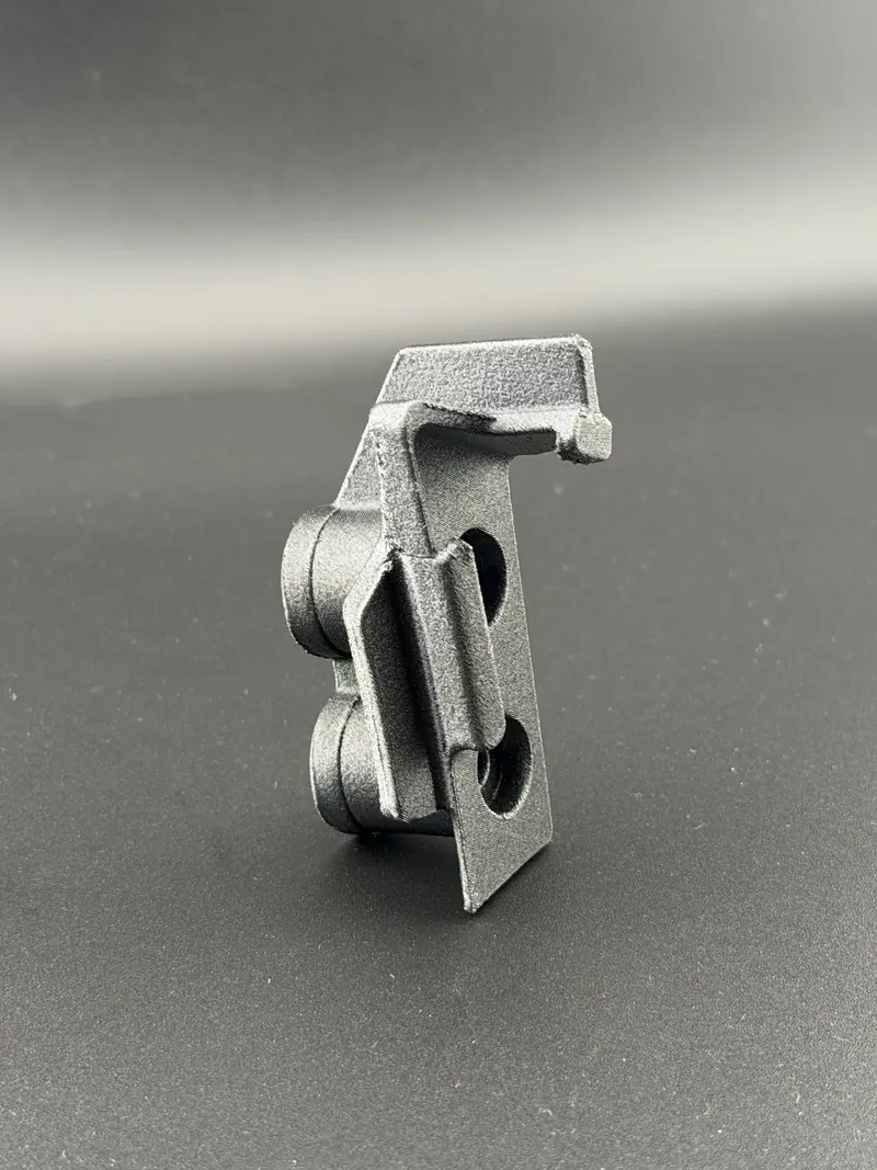 Black 3D printed bracket standing on end: a flat tab with two round holes, two round bosses on one side and a hooked arm at the top.