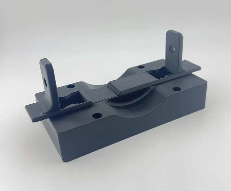 Dark blue-gray 3D printed fixture block with a curved cutout across its top, four round holes, and a flat strip with two upright tabs that each have a hole.