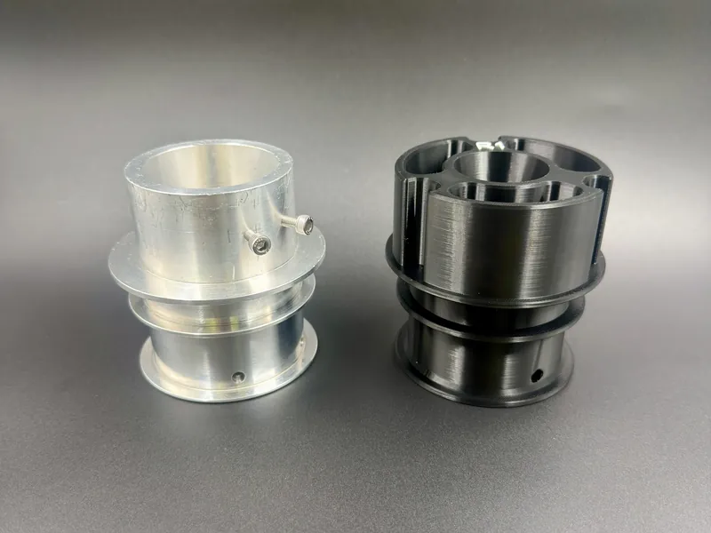 A machined aluminum wire hub with two socket-head screws next to its redesigned black 3D printed alternative, both stepped hubs with flanges.
