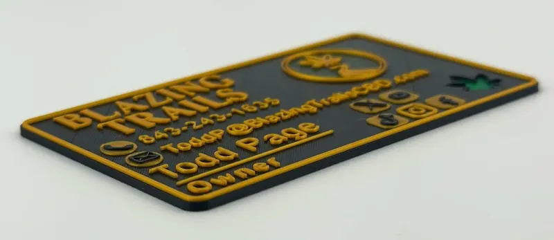 3D printed business card for Blazing Trails with raised gold lettering on black, listing owner Todd Page, phone, email and social icons beside a circular cannabis-leaf logo.
