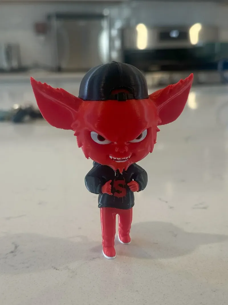 A figurine printed by us: a red devil character with large pointed ears, wearing a backwards black cap and a black hoodie, standing on a countertop.
