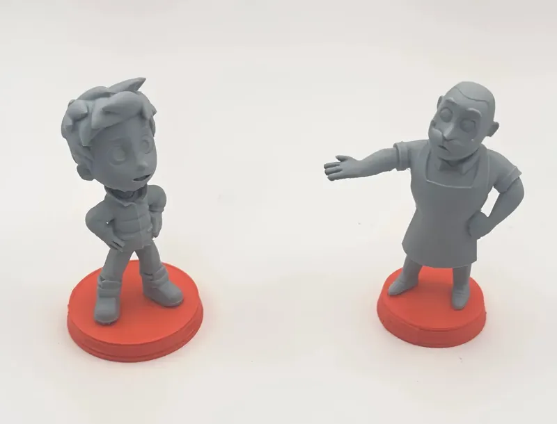 Two gray character figurines on orange bases, printed by us in resin: a young boy with tousled hair, hands on his hips, facing an older man with a mustache and apron gesturing toward him.