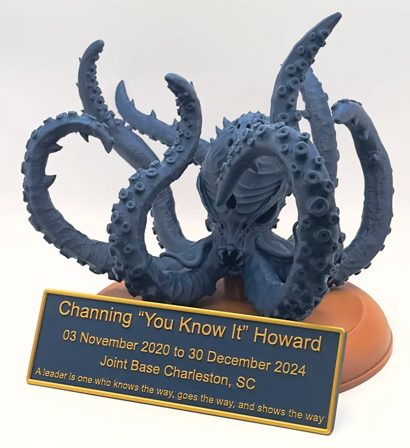 A sculpture printed by us: a dark blue Cthulhu-style figure with a horned skull face and coiling tentacles on an orange base, mounted with a gold-lettered farewell plaque for Joint Base Charleston, SC.