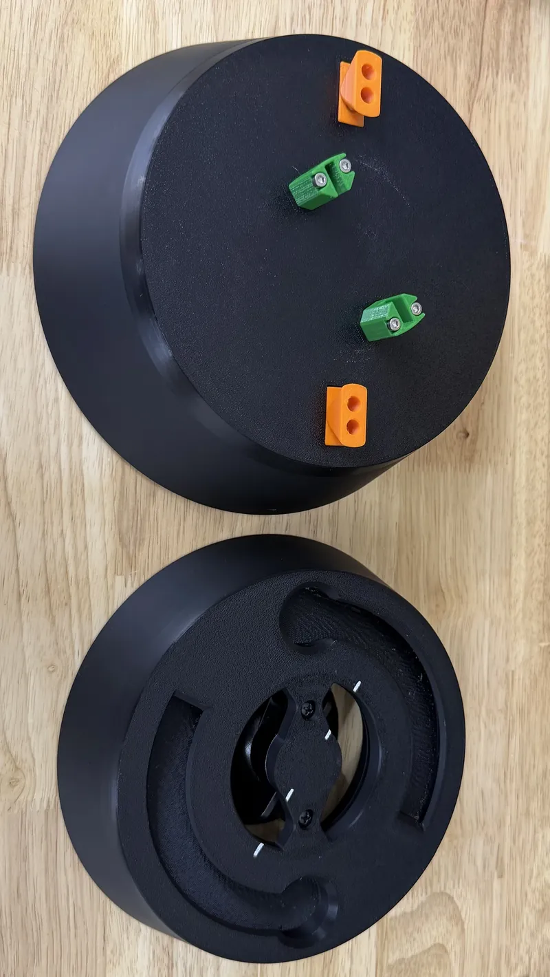 Two black round parts on a wooden table: one face up with two orange two-hole blocks and two green clamp blocks screwed to it, the other turned over to show its hollow inside, with curved slots and a center fitting.
