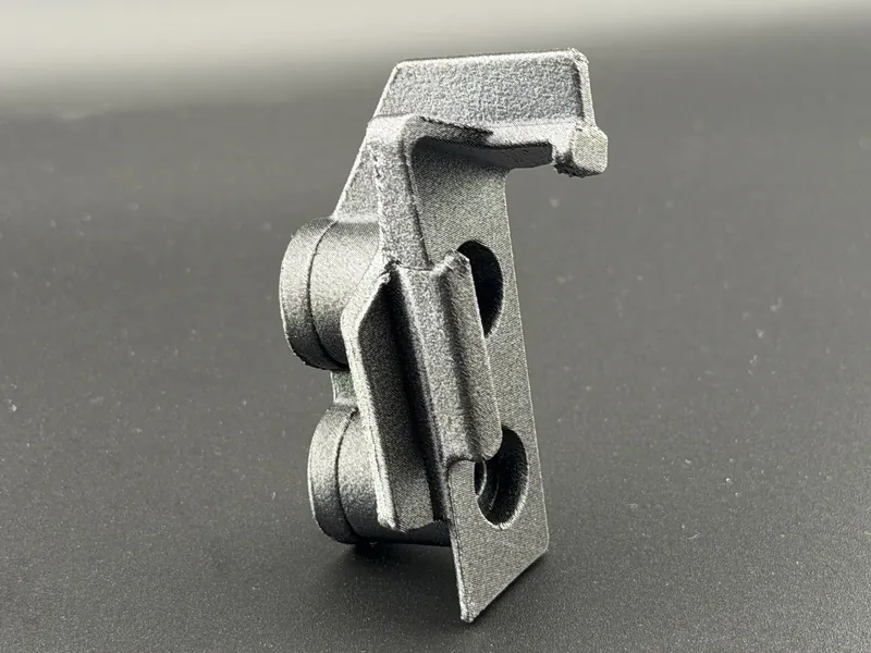 Small gray 3D printed spring-latch clip with two roller bosses and a mounting tab, shown close up on a dark background.